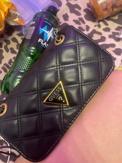 A black quilted clutch purse with a gold Guess logo is resting on a person's leg. To the left of the purse is a green bottle with the word "FLASH" written on it. The background consists of a leopard-print fabric and some other objects that are out of focus, possibly snacks. The scene appears to be indoors, and the lighting suggests it might be evening or a dimly lit room. There is no indication of the city of Shcheglovo, Russia.