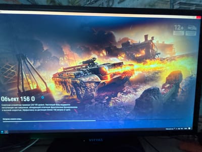 The image shows a computer screen displaying a loading screen for a video game. The game appears to be a tank combat simulation, with a prominent image of a tank in the foreground, engulfed in flames and smoke, with a train in the background also on fire. The scene is dramatic and action-oriented, set in a war-torn or industrial environment.
Visible text on the screen includes game information in Russian, such as "Объект 156 0" (Object 156 0), which is described as a Soviet premium tank destroyer of Tier VIII, effective in supporting offensive operations with excellent frontal armor and high speed, particularly at ranges under 150 meters. The text "Загрузка клиента игры...." (Loading game client....) indicates that the game is in the process of loading. Age rating icons, including "12+", are also visible.
In the lower right corner of the screen, system information is displayed, including "Рабочий стол" (Desktop), an icon indicating audio, and the time "10:06". The brand name "VITYAS" is visible on the monitor bezel.
The overall impression is one of anticipation for intense gameplay, with the visuals and text suggesting a focus on armored warfare and strategic combat. The lighting and effects create a sense of chaos and destruction.