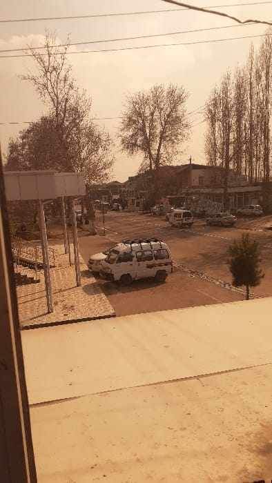 A street scene shows several vehicles parked along the side of a road, including a white van. Bare trees line the street, and a building borders the right side of the frame. The image has a muted, sepia-toned color scheme, giving it an older or filtered appearance. The scene is viewed from an elevated position, possibly from a window or balcony.