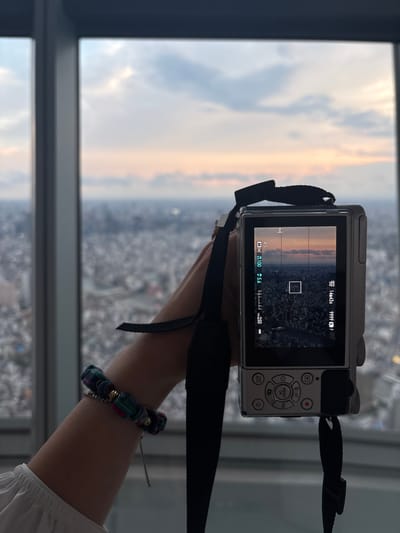 An image taken from a high vantage point in Sumida, Japan, shows a person's arm holding a silver digital camera in the foreground. The camera's LCD screen displays a cityscape at sunset, with grid lines overlaid, indicating a photographic composition. The person is wearing a white, ruffled shirt and a colorful beaded bracelet on their wrist. Beyond the arm and camera, a blurred panoramic view of a dense urban environment stretches to the horizon under a sky transitioning from light blue to soft orange hues, suggesting either dawn or dusk. The window frame of the building is visible on the left side of the image.