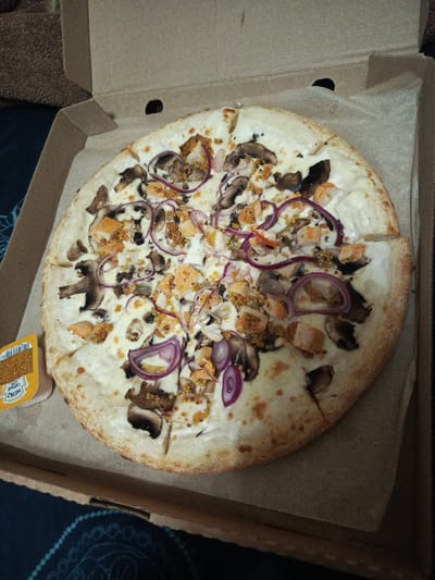 A whole, round pizza with white sauce and toppings of sliced mushrooms, red onion rings, and chunks of fish or chicken is presented in a cardboard pizza box. A small yellow packet of Heinz mustard is visible on the left side of the pizza. The pizza is cut into eight slices, with one slice slightly askew. The background of the image consists of the brown cardboard box and a dark blue fabric with a light blue circular pattern. The image appears to be taken in an indoor setting, possibly a home, and does not provide any specific details about the city of Salavat, Russia.