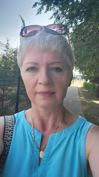 A woman with short, light-colored hair and sunglasses perched on her head is taking a selfie outdoors. She is wearing a sleeveless blue top and a gold necklace with a pendant. The background shows a paved walkway lined with trees and a fence. A person is walking in the distance. The scene appears to be in the city of Belaazyorsk, Belarus.