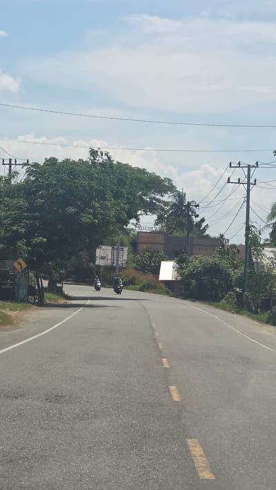 The image depicts a road in Baroh Musa, Indonesia, with two motorcycles traveling on it. The road is gray and has broken yellow lines marking the lanes. A curve in the road is visible in the foreground. Utility poles with overhead wires run along the side of the road. Lush green vegetation lines the roadside, including trees and bushes. A building with a sign is partially visible behind the trees. The sky is blue with scattered white clouds.