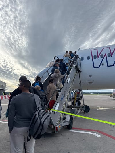 In Künersberg, Germany, on an overcast day, a group of passengers is boarding an airplane via a mobile stairway. The airplane, a white Boeing with a distinctive purple and pink "W" logo on its tail, is parked on the tarmac. The stairway, labeled "ALLGATE," is silver and has a yellow safety tape stretched across its base, delineating a boundary on the concrete surface. Several people, dressed in casual clothing, are ascending the stairs, some carrying backpacks and bags. The sky above is filled with dramatic, textured clouds. In the background, airport infrastructure is visible, including a building with red and white barriers.