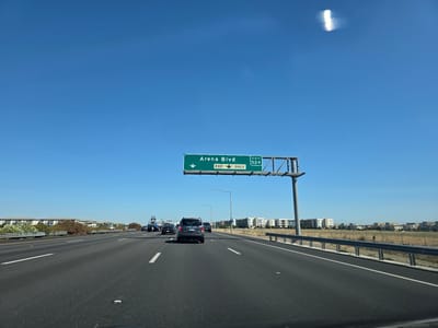 A clear blue sky with a lens flare anomaly is visible above a multi-lane highway in Sacramento, California. A green overhead highway sign indicates "Arena Blvd" and "EXIT 524" with an arrow pointing down and to the left for the exit. Several cars are traveling on the highway, including a dark gray SUV in the foreground. The surrounding environment features low-rise residential buildings and dry grass fields. The perspective suggests the image was taken from inside a moving vehicle.