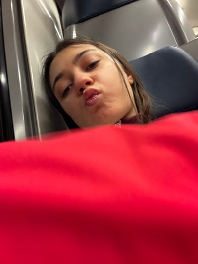 A person with brown hair and pouting lips lies against a gray surface next to a blue upholstered seat. The individual's face is turned slightly toward the camera, with eyes gazing downwards. A large, blurred red shape dominates the right side of the image, partially obscuring the background and figure. The setting appears to be inside a train or other form of public transport.