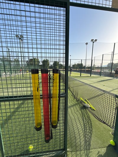 The image shows an outdoor padel tennis court in Vidreres, Spain. The court is made of green artificial turf and is enclosed by a metal fence. A net divides the court in half. Three transparent tubes, two yellow and one red, filled with padel balls are attached to the fence on the left side of the image. Several yellow padel balls are visible on the court. In the background, there are more padel courts, trees, and a clear blue sky with the sun setting, casting long shadows across the court. Two people are visible on the courts; one is standing near the net on the right, and another is playing on a court further in the background. There are also some stadium lights visible above the courts.