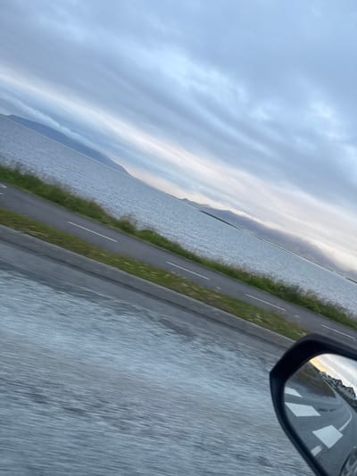 The image depicts a view from a vehicle in Seltjarnarnesbær, Iceland. A body of water stretches across the frame, bordered by a road with a white line marking. Beyond the water, mountains are visible under a cloudy sky. A side-view mirror of the vehicle reflects a street with buildings and road markings. The overall lighting suggests it is either early morning or late evening. The scene is characterized by a sense of movement, as indicated by the blurred foreground.