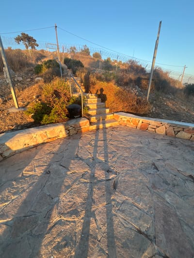 The image presents an outdoor scene in Kafr Bara, Israel, under a clear blue sky, illuminated by low-angle golden sunlight casting long shadows. In the foreground, an irregularly paved stone patio with varying shades of gray and brown flagstones extends. A low, curved stone retaining wall composed of rough-hewn stones defines the right edge of this patio. Two distinct, elongated human shadows are cast prominently across the patio and onto the steps beyond; one shadow suggests an individual holding a mobile device.

Mid-ground features a set of stone steps with a silver-colored metal handrail, ascending a moderately steep, arid hillside. To the immediate left of the steps, a black cylindrical trash receptacle is situated, bearing Hebrew text "כפר ברא" (Kafr Bara). The hillside is covered with dry, sparse vegetation, including bushes, some green foliage near the steps, and scattered rocks. A utility fence, constructed from metal posts and multiple strands of barbed or razor wire, runs horizontally across the mid-ground, stretching from left to right across the incline. In the background, additional dry vegetation and a few scattered trees are visible on the hill's crest.