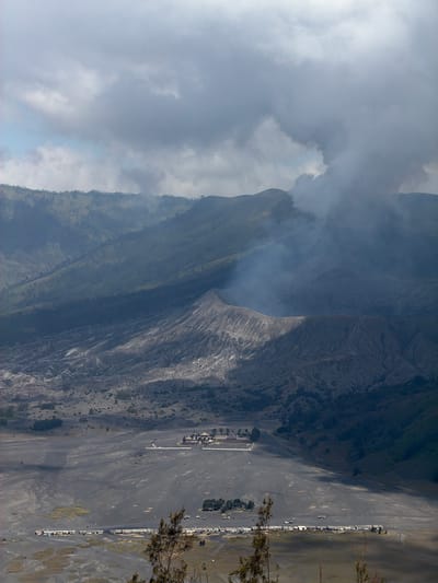 The image captures a panoramic view of Mount Bromo, a volcano in East Java, Indonesia, specifically looking towards Cemorolawang. In the foreground, a vast expanse of dark volcanic sand stretches out, punctuated by patches of dry grass and sparse vegetation. Several structures are visible in the distance; a long, low building complex with numerous small stalls or shops lines the bottom left of the frame, some with umbrellas erected outside. Further back, near the center of the image, a more substantial, multi-building compound with red-tiled roofs sits on the volcanic plain.
The middle ground is dominated by the rugged, ashen slopes of Mount Bromo, with its caldera clearly visible. Wisps of white smoke or steam emanate from the volcano's crater, indicating ongoing geothermal activity. The background is comprised of rolling, green hills and mountains, their slopes covered in vegetation and trees. The sky above is partly cloudy, with dramatic grey clouds casting shadows across the landscape. The overall impression is one of raw, natural power and a stark, volcanic environment.