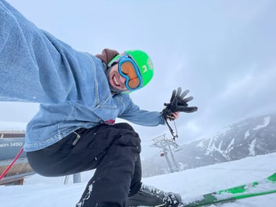 A person is captured in a dynamic, low-angle selfie on a snow-covered ski slope in Mušovića Rijeka, Montenegro. The individual is dressed in a light blue denim jacket over a brown hooded garment, black ski pants, black ski boots, and green skis. A bright green ski helmet with orange goggles covers their head. The person's mouth is open in an expression consistent with excitement or exertion, and their right hand, clad in a black ski glove, is raised with fingers splayed. To the left, a wooden structure, possibly a ski lift station, displays a partial inscription "sin 1450" and a red rope. In the mid-ground, multiple ski lift towers equipped with red chairlifts ascend a snow-covered mountain. The background consists of distant, snow-covered mountainous terrain under an overcast, light grey sky.