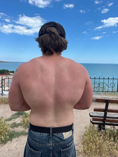 The image shows a person from behind, facing the sea. They are wearing a dark baseball cap and blue jeans. The person's back and shoulders are exposed, revealing tanned skin. In the background, there is a body of water, possibly the Black Sea, with a clear blue sky dotted with white clouds. A black metal fence with decorative elements runs horizontally across the middle ground, separating the viewer from a beach area with scattered people and structures. A wooden bench is visible on the right side of the frame. The terrain in the foreground is dry and gravelly, with patches of dry grass. The location is identified as Kryzhanivka, Ukraine.