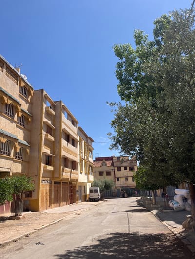 The image depicts a street scene in El Hajeb, Morocco, under a clear blue sky. On the left, a multi-story building with a textured, light brown facade and decorative green tiled roof accents lines the street. The building features multiple windows with brown shutters and arched window frames. Opposite this building, a road curves gently into the distance.
Several trees are visible, including a prominent olive tree on the right, its branches casting dappled shadows on the road. A smaller, leafy green tree stands near the building on the left.
A white van is parked on the right side of the road, further down. A few individuals can be seen in the distance, near some market stalls. On the right sidewalk, large white sacks are stacked. The street surface is asphalt, showing signs of wear, and the sidewalks are a mix of concrete and dirt. The overall impression is a residential or commercial street in a Moroccan town on a sunny day.
