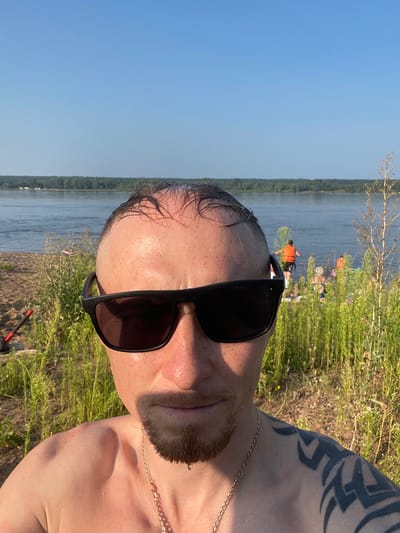 A medium shot selfie taken outdoors on a sunny day shows a man in the foreground. The man is facing the camera and wearing black sunglasses. He has a shaved head, a goatee, and a tribal tattoo on his right shoulder. A gold chain is visible around his neck. Behind the man, a body of water, possibly a river, stretches across the background, with a tree-lined shore in the distance. Several people are visible in the water and on the shore in the middle ground, some wearing orange life vests. The foreground is occupied by tall green grass and a sandy area. The scene appears to be a recreational area near a body of water, consistent with the location of Соколовка, Russia, which is known for its natural landscapes and proximity to the Oka River.