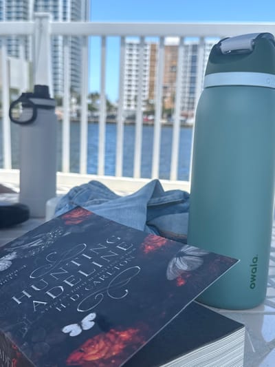 The image captures an outdoor scene on a sunny day in Hallandale Beach, United States. In the foreground, a paperback book titled "Hunting Adeline" by H.D. Carlton rests on a light-colored, possibly tiled, outdoor surface. A teal Owala-branded water bottle stands upright to the book's right. Folded light-wash denim material, a partially obscured light grey water bottle, and a dark, indistinct object are also present on the surface. Beyond these items, a white railing fronts a body of water, likely an intercoastal waterway or canal. Multiple high-rise residential buildings are visible across the water, set against a clear blue sky. No individuals are present.