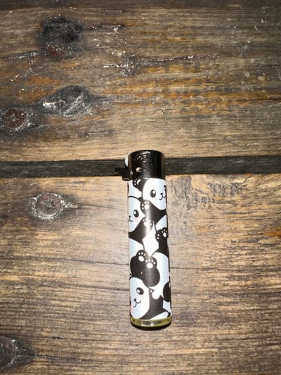 A black and white patterned lighter lies horizontally across two weathered, dark wood planks. The lighter features a repeating panda face design. The left end of the lighter has a shiny, black metallic finish. Visible screws are embedded in the wood near the lighter's position. The wood planks exhibit a rustic texture and a noticeable seam where they meet. Dark patches and imperfections are scattered across the wood surface.