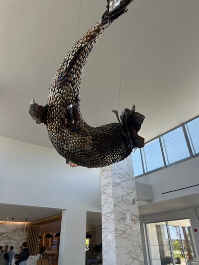 A large metallic sculpture of a stylized fish hangs from the ceiling in a bright, modern interior. The fish is constructed from numerous small, round metal pieces resembling scales. It has a prominent head with a decorative, leafy element and fins. The interior features white walls, skylights, and windows. A marble-patterned wall is visible, as is an open doorway leading to a restaurant or cafe area with people and tables. Several people are visible in the lower right of the frame. This appears to be the Ikos Oceania Hotel, Kalyves Polygyrou, Greece.