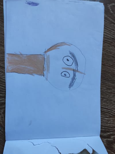 A child's drawing on white paper depicts a simplified human face with two oval eyes and a horizontal line for a mouth. The face is within a roughly circular outline. To the left of the face is a rectangular shape colored in orange, representing a body or neck. Above the face, to the left, are some scribbled purple lines. The drawing is lying on a wooden surface, likely a table, with visible wood grain. The image is oriented sideways. There is no indication of the city of Kipshek, Russia, in the provided image.