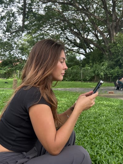 In Tambon San Phi Suea, Thailand, a young woman with long, wavy brown hair is captured from the side, looking down at her smartphone. She is wearing a black t-shirt and dark gray pants, seated outdoors on a patch of vibrant green grass. Her left arm rests on her lap, and her right hand holds the phone. Behind her, a large tree with an intricate canopy of branches and leaves dominates the background. In the distance, a blurred scene shows two people sitting at a table with chairs. The environment suggests a park or outdoor recreational area.