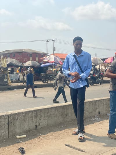 The image presents an outdoor street and market scene in Awjaw, Nigeria, under a partly cloudy sky. In the foreground, a Black male stands on a dirt verge adjacent to a concrete median, facing slightly left and looking towards the camera. He wears a light blue long-sleeved collared shirt, black trousers, and two-strap sandals. A black cross-body bag is slung over his right shoulder, and white wired earphones are visible around his neck. His hands are interlocked at waist height, displaying multiple rings. To his immediate right, a second individual is partially visible, wearing a grey t-shirt, blue jeans, and light brown slip-on shoes.
Behind the concrete median, a paved asphalt road extends into the mid-ground. Two other Black males are observed walking from left to right across the road; one wears a dark blue long-sleeved shirt and brown trousers, while the other wears a light-colored patterned short-sleeved shirt and dark trousers. The far side of the street is occupied by numerous market stalls, sheltered by various large umbrellas, including black and red-white-blue striped designs. Merchandise, including stacked items and bottles, is visible on tables, and motorbikes are parked among the stalls. A white banner featuring a portrait is attached to a stall on the far left.
In the background, a two-story red-painted building with a dark roof stands prominently on the left, next to other structures. Overhead utility wires are suspended between poles against the sky. On the dirt verge in the immediate foreground, a white cylindrical object and a dark, irregular object are lying on the ground.