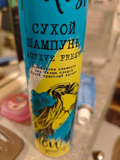 The image shows a close-up of a blue and yellow aerosol can, likely a dry shampoo product, placed on a surface. The can features prominent text in Russian and English. The Russian text reads "СУХОЙ ШАМПУНЬ," which translates to "DRY SHAMPOO." Below this, the English text "ACTIVE FRESH" is visible, along with smaller Russian text that says "Длительная свежесть. Без белых следов. Яркий чудесный аромат," meaning "Long-lasting freshness. No white residue. Bright wonderful aroma." A stylized illustration of a woman's head with long, wavy hair adorned with blue flowers is printed on the can. At the bottom of the can, there appears to be a logo with stylized text, possibly the brand name, with "NEW" in large letters above it. The can is positioned at an angle, with the background blurred and out of focus, showing hints of other objects on what appears to be a countertop or shelf. The lighting suggests an indoor setting, possibly a bathroom or a dressing area. The overall impression is a product shot focusing on the design and key features of the aerosol can. The location context provided is Berdsk municipality, Russia.