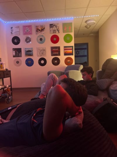 Two people rest on a couch. One is visible, face down, while the other is slightly behind. A wall displays an array of records, illuminated by a string of lights. In the background, a doorway reveals a partially visible Israeli flag. Miscellaneous items are scattered on the floor near a small, black shelving unit. The room's lighting is warm.