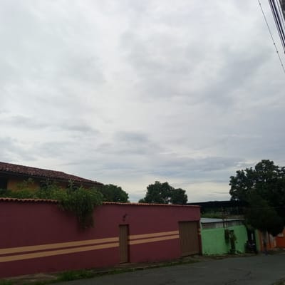 The image depicts a street scene in El Limón, Venezuela, under an overcast sky. In the foreground, a dark asphalt road is visible, with patches of grass along its edge. A prominent, long, deep red wall dominates the midground, adorned with two horizontal pale yellow stripes. This wall features a wooden gate on the right side and a smaller wooden door on the left, with the house number "82" above it. Behind the red wall, a portion of a house with a terracotta tile roof and yellow siding is visible, partially obscured by lush green foliage.
To the right of the red wall, a bright green wall extends, also with a corrugated metal roof structure behind it. This green section appears to be part of another property or dwelling. Further to the right, a darker colored building with a metal roof and fencing is seen, and a few individuals are present near the entrance. The background consists of dense green trees and vegetation, suggesting a residential area with abundant plant life. Overhead, several dark power lines run diagonally across the sky on the right side of the frame.