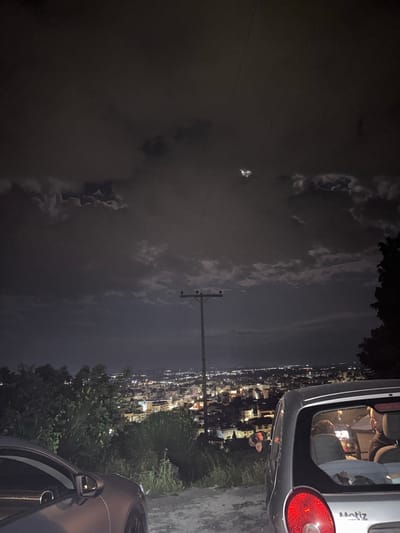 The image depicts a night scene in Xanthi, Greece, overlooking the city. The sky is dark with scattered clouds, and a full moon is visible. A utility pole stands prominently in the foreground, with power lines stretching across the sky. Two cars are parked on the side of what appears to be a road or a viewing point. The city lights illuminate the landscape below, with buildings and streets clearly visible. Inside the trunk of the silver car there appears to be three individuals. There are trees to the bottom and right of the image.