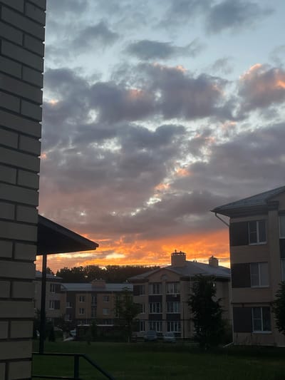 This is a photograph of a sky filled with dramatic clouds at sunset, as seen from within a residential complex in Russia. The clouds are a mix of dark grays and lighter whites, with streaks of vibrant orange and pink where the setting sun is breaking through. The light from the sunset illuminates the edges of the clouds and casts a warm glow on the scene.
In the foreground, a brick fence is visible, with a portion of a dark, angular structure, possibly part of a building or a piece of equipment, protruding from the bottom edge. Behind the fence and to the right, apartment buildings with multiple stories and numerous windows are visible. They appear to be modern residential structures. Trees and green grass are also present, suggesting a well-maintained outdoor area within the complex. The overall impression is a residential setting during a visually striking sunset. The time of day is clearly sunset due to the colorful sky. The weather appears to be partly cloudy, creating a dynamic and colorful sky. There is no visible text in the image.