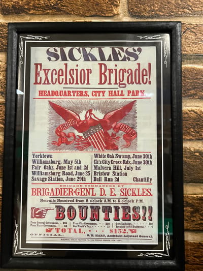 A framed vintage poster advertises Sickles' Excelsior Brigade, headquartered at City Hall Park. It lists various battles, including Yorktown, Williamsburg, Fair Oaks, and Bull Run 2nd. The brigade is commanded by Brigadier-General D.E. Sickles. Recruits were received from 8 a.m. to 6 p.m. The poster prominently displays "Bounties," with monetary amounts listed from general, state, and city governments, along with corn exchange and premium to old regiments, totaling $152. The official is O.H. Hart, Assistant Adjutant General, with printing by Baldwin, Stein Printer, NYC.