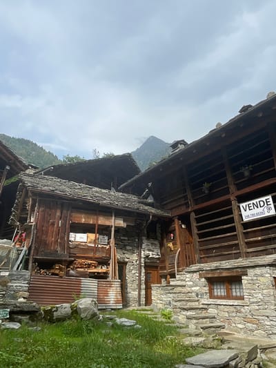 The image depicts a rustic, multi-story building constructed from stone and dark wood, with slate roofs. The architecture suggests a historical dwelling or farmstead, typical of mountainous regions. The building is set against a backdrop of a cloudy sky and a distant mountain peak.
In the foreground, there is overgrown green grass and several large grey rocks. The building itself shows signs of age and use. The lower section appears to be made of rough-hewn stone, with a wooden door and a small window with a lattice. A stone staircase leads up to a wooden entrance. Above the stone base, the structure transitions to weathered wood, with stacked firewood visible in an open section.
A prominent feature is a "VENDE" (for sale) sign with a phone number, indicating that the property is on the market. There are also two small potted plants placed on shelves in the wooden structure, adding a touch of color. The overall impression is one of a traditional, possibly abandoned, property in a rural, mountainous setting.
While the image is not definitively identifiable as being in Alagna Valsesia, Italy, the architectural style of stone and wood buildings with slate roofs is characteristic of many villages in the Italian Alps, including Alagna Valsesia, which is known for its Walser heritage and well-preserved traditional homes.