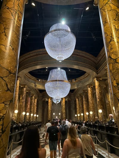 The image depicts the interior of a grand, ornate hall, likely a themed attraction. Dominating the scene are two massive, tiered chandeliers, each adorned with countless crystal-like droplets that reflect light. The hall features numerous tall, fluted columns with a marble-like finish in shades of gold and brown, creating a sense of opulence. A circular architectural element frames the upper portions of the hall, with more decorative molding.
In the foreground, a group of people are walking away from the camera, observing the hall. Their clothing suggests a casual setting. The people are positioned on a tiled floor with a checkered pattern in white and grey. Along the sides of the hall, long counters with individual desks or workstations are visible, each illuminated by a spherical lamp. Some figures, possibly animatronics or mannequins dressed as clerks, are seated at these stations. Ropes on stanchions delineate pathways for visitors. The overall atmosphere is dimly lit, with the chandeliers and lamps being the primary light sources. The location is described as Three Rivers, United Kingdom.