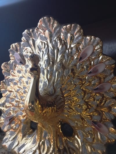 A decorative figurine, identified as a peacock, is centrally depicted against a dark, out-of-focus background. The object is predominantly gold-colored, exhibiting a highly reflective and polished surface. The fanned tail structure features intricate sculpted patterns, partially coated with silver glitter. Embedded within the tail are multiple ovular, shell-like embellishments, each displaying a dark, spiral pattern against a lighter, opaque base. The peacock's body and neck are smooth and maintain the gold finish. No human subjects, animals, or active interactions are present in the image. The object is situated in Nasiriyah, Iraq.