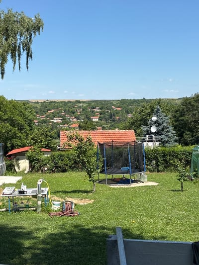 In Pisanets, Bulgaria, a backyard scene unfolds under a clear blue sky. A large trampoline with a blue safety net is centrally located on a circular concrete pad. To the left of the trampoline, a temporary sink setup is arranged on a blue-legged table. This setup includes a basin, a faucet with a yellow hose connected, several bottles of liquid, and a dish rack. A coiled red garden hose lies on the grass nearby. In the background, a village with red-tiled roofs is nestled among green, rolling hills covered in trees. A tall, blue-green conifer tree stands to the right of the trampoline, with satellite dishes mounted on a white structure behind it. Overhanging branches of a leafy tree frame the upper left corner of the image. The foreground is a well-maintained lawn with scattered white clover flowers.