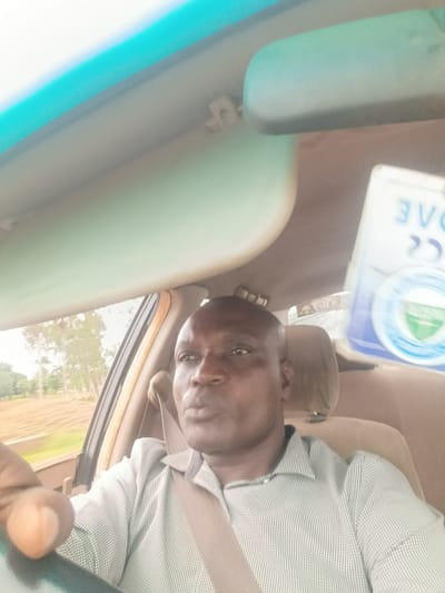 A selfie shows a dark-skinned man driving a car. He is wearing a light-colored, pinstriped button-down shirt and a seatbelt. The man is looking towards the camera with his mouth slightly open. The interior of the car is visible, including the dashboard, rearview mirror, and a car air freshener. Outside the car, trees and fields can be seen. The overall lighting suggests it is daytime. The man appears to be in the city of Ganawuri, Nigeria.
