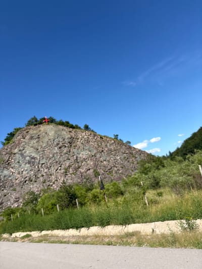 An outdoor, eye-level shot captures a rocky hill topped with trees and a bright red cross under a clear blue sky in Malo Buchino, Bulgaria. The hill is covered in sparse vegetation and rough, grey rock formations. A road is visible in the foreground, with a grassy verge and a low stone wall separating it from the base of the hill. The hill itself is steep and appears to be made of a rough, grey, and greenish stone. A cluster of green trees crowns the summit, surrounding the prominent red cross. The sky is a vibrant, cloudless blue, with only a few wisps of white clouds in the upper right corner. The overall scene is serene and rural, with the cross serving as a striking focal point on the landscape.