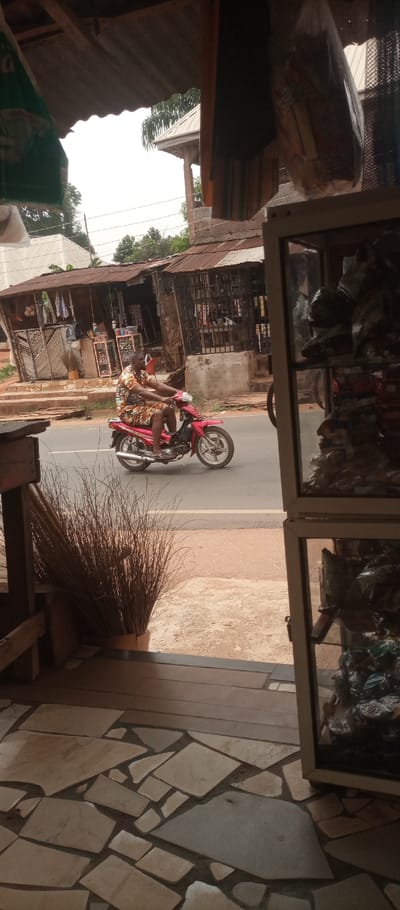 The scene depicts a street view in Umuoji, Nigeria, observed from the interior of a commercial establishment. In the immediate foreground, a floor is visible with irregularly shaped light brown and beige tiles. To the left, a bundle of dried straw brooms stands upright next to a dark wooden structure. To the right, a white-framed glass display cabinet contains numerous small items packaged in clear plastic bags. The overhead view shows the underside of a corrugated metal roof and various items suspended, including a green bag on the left and a clear plastic bag on the right.
Across the dirt shoulder, an asphalt road traverses the midground. A male individual, wearing a short-sleeved patterned shirt and patterned shorts with sandals, is operating a red motor scooter (moped style) and moving from left to right along the road.
In the background, multiple single-story structures with rusted corrugated metal roofs line the opposite side of the street, appearing to be small shops or market stalls displaying merchandise. One structure on the right features a barred window or entrance. Further back on the left, a building with a lighter-colored facade and a slanted roof is partially visible. Dense green foliage and trees are present behind the background structures, under a bright, overcast sky.