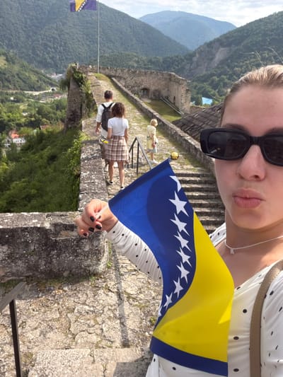 This media file captures a selfie of a young woman holding the flag of Bosnia and Herzegovina. She is wearing sunglasses and a white dress with black polka dots. Her left hand, with black nail polish, is visible in the foreground, holding the flag.
Behind her, a historic stone fortress with crenellated walls dominates the scene. The fortress appears to be situated on a hilltop, overlooking a lush, green valley with a town nestled within it. The terrain is mountainous, with misty peaks visible in the distance, suggesting a cloudy or hazy day.
Several other individuals are present within the fortress, appearing to be tourists exploring the site. One person in a white shirt and plaid skirt is walking away from the camera, while another person in a white shirt with a backpack is also seen further down the stone pathway. A faint glimpse of a blue and yellow flag, similar to the one being held, is visible on a pole in the distance on the left.
The overall impression is one of tourism and cultural exploration in a historical setting. The lighting suggests it is daytime. The location cues and the flag strongly indicate the scene is Jajce, Bosnia and Herzegovina. There is no visible text in the media file.