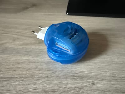 A blue electric mosquito repellent plug sits on a light-colored, wood-grained surface. The device has two prongs extending from a white plastic adapter. The blue plastic housing is oval-shaped with vents and a slot where a repellent insert is visible. A small red indicator light is present on the top of the device. The background shows a dark, out-of-focus object, possibly a monitor. The surface the item rests on appears to be a table or desk. The scene is indoors. This image does not contain information that allows for the specific location within Khazar Raion, Azerbaijan.