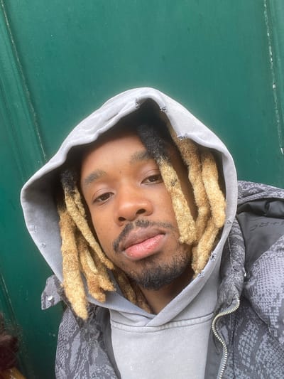 A man with dreadlocks lies in a hooded jacket on a dark green surface. The man's head is positioned toward the top left of the frame. He has brown skin, and his dreadlocks are blonde with dark roots. His beard is dark and slightly unkempt. The hood of his gray jacket frames his face. Part of a gray jacket with a snakeskin pattern is also visible. The location cannot be determined based on visual cues in the image.