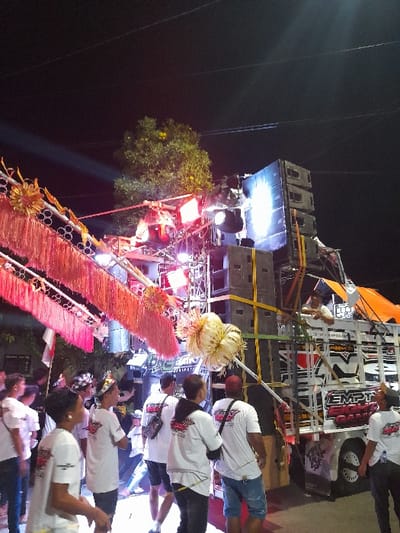 This image captures an outdoor nighttime scene in Kedungbanteng, Indonesia. The central focus is a large sound and lighting system. On the right, a white flatbed truck, marked "EMPTY EIGHTY GALA," supports a tall stack of dark speaker cabinets. Adjacent to and above the truck, a metal truss structure holds numerous stage lights emitting red, blue, and white light, with one bright white beam projecting upwards into the dark sky.

On the left, another structure is adorned with multiple reddish-orange, tassel-like decorative strips. Two large, spherical, woven ornaments of a light, yellowish color are also suspended, one prominently in the mid-ground. A green-foliaged tree is visible behind the decorative elements on the left, illuminated by the stage lights. Overhead utility wires traverse the dark sky.

Approximately 10-15 individuals, predominantly adult males, are visible in the foreground and mid-ground. Most are wearing identical white short-sleeved t-shirts featuring a red graphic design on the back. Some individuals wear shorts, others dark pants. Their stances suggest observation of the equipment setup or anticipation. One individual is positioned on top of the white truck, near the sound equipment. The ground beneath the individuals appears paved.