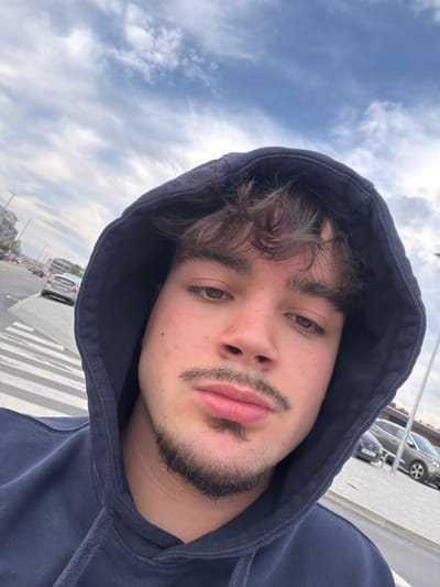 A young male, appearing to be in his late teens or early twenties, is depicted in a close-up, head-and-shoulders portrait shot, likely a selfie. He has brown, wavy hair partially covered by a dark blue hooded sweatshirt, which he is wearing with the hood up. His facial features include brown eyes, a mustache, and a goatee. His expression is neutral.
The background reveals an outdoor urban or suburban setting. The sky above is blue with scattered white clouds. To the left and behind the subject, a paved road or street is visible, featuring a white-striped pedestrian crossing in the immediate mid-ground. Several vehicles are present on the road and in a visible parking area to the right. These vehicles include a light-colored sedan and multiple darker cars. Structures consistent with buildings or commercial establishments line the street in the distance. A utility pole is partially visible on the right side of the frame. The scene is situated in the city of Felgueiras, Portugal.