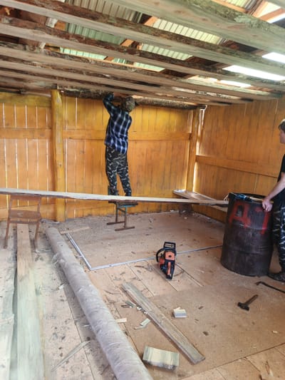 A person wearing a blue and black plaid shirt and camouflage pants is working on the ceiling of a wooden structure. The person appears to be installing wooden planks. The environment is rustic, with exposed wooden beams and unfinished walls. Sawdust and construction debris are scattered on the floor. A chainsaw, a barrel, and a hammer are visible on the ground. Another person, wearing a black t-shirt and camouflage pants, stands to the right of the frame, observing. The scene suggests ongoing construction or renovation work.