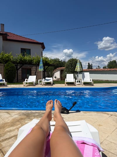 The image depicts a person relaxing by a swimming pool on a sunny day in Malko Gradishte, Bulgaria. The foreground shows the person's legs and feet resting on a white lounge chair, with a pink and white striped towel draped over the chair. The swimming pool, filled with vibrant blue water, occupies a significant portion of the right side of the image. The pool area is surrounded by a light-colored tiled deck.

In the background, there are several empty white lounge chairs and a colorful umbrella. A white building with a red-tiled roof is visible to the left, with green foliage and trees surrounding it. The sky is a clear, bright blue with scattered white clouds. The presence of power lines suggests an outdoor residential or recreational area. The overall mood is one of relaxation and enjoyment of a warm, sunny day. No visible text is present in the image.
