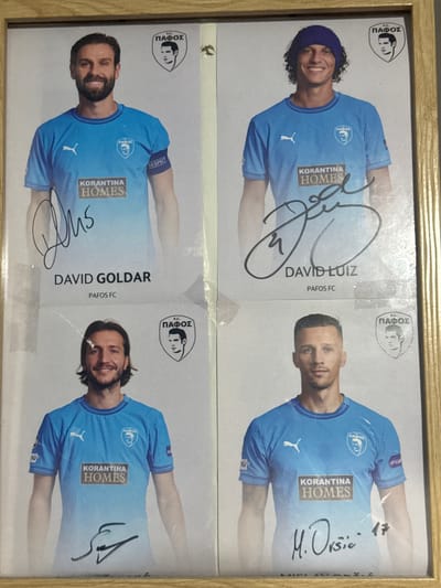 The image displays a framed collection of four football player cards, arranged in a 2x2 grid. Each card features a portrait of a male athlete in a light blue football jersey, a team logo, and a sponsor's logo. All four players are associated with "Pafos FC" and have signatures on their respective cards. The background of each card is a plain light gray.
The top row shows two players. The left card features a player with short dark hair and a beard, wearing a blue jersey with a white Puma logo and a crest that reads "FC ΠΑΦΟΣ". The right card shows a player with curly dark hair and a purple headband, also wearing a similar blue jersey with the "FC ΠΑΦΟΣ" crest.
The bottom row also features two players. The left card depicts a player with medium-length dark hair and a beard, in a blue jersey. The right card shows a player with short blond hair, also in a blue jersey. Both bottom row cards have the sponsor's logo "KORANTINA HOMES" prominently displayed on the chest. The player on the right has a handwritten number "17" next to his signature.
The overall presentation suggests a promotional item or collectible related to Pafos FC. The image does not provide any specific visual cues to determine its exact location within the city of Yeroskipou, Cyprus, other than the team's affiliation.