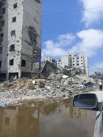 In Jabalia, Palestinian Territories, a scene of significant destruction is depicted. In the foreground, a partially submerged car's side mirror reflects a brown, muddy puddle that appears to be mixed with debris. The puddle reflects the image of a damaged building. Adjacent to the puddle is a large pile of rubble, consisting of concrete, rebar, and various forms of refuse, including plastic bags and bottles.
Dominating the left side of the image is a tall, multi-story building that has sustained severe damage. Its concrete facade is riddled with numerous holes, and large sections of the structure are collapsed, revealing exposed interiors and broken floors. Debris from this building has fallen and contributed to the pile of rubble in the foreground.
Further in the background, more buildings are visible, also exhibiting signs of damage, though to a lesser extent than the prominent structure on the left. Some of these buildings appear intact from a distance, while others show visible damage. The sky above is blue with scattered white clouds. A single individual, dressed in a white shirt and dark pants, is seen walking away from the camera on the right side of the frame, near the edge of the debris field. The overall impression is one of widespread destruction and the aftermath of conflict.