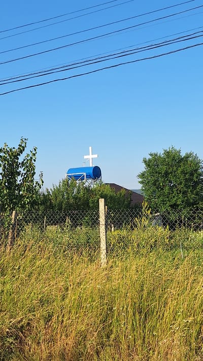 The image shows a rural scene with a water tower topped by a white cross, set against a clear blue sky. The water tower is a large, cylindrical blue tank supported by a metal structure. The cross is white and prominently placed on top of the tank. In the foreground, a chain-link fence runs horizontally across the frame, separating the viewer from a grassy field with tall, dry grass and some scattered green vegetation. Behind the fence and the water tower, there are trees and what appears to be a building with a dark roof. Overhead, several power lines are visible against the sky. The scene is likely located in a village or rural area, given the presence of the water tower, the fence, and the vegetation. Sărata-Galbenă is a village in Moldova, which aligns with the appearance of the scene. There are no people visible in the image.