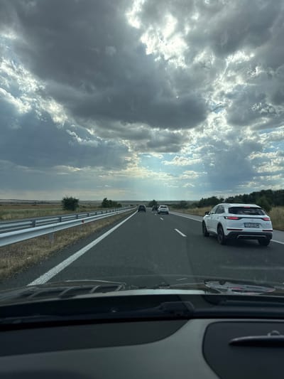 The image depicts a view from inside a vehicle driving on a highway. The sky is filled with dramatic, dark clouds, with patches of sunlight breaking through in the distance, creating visible sunbeams. The highway is asphalt with white lane markings. A silver guardrail runs along the left side of the road, separating it from a dry, grassy embankment. In the distance, the landscape is flat and appears to be agricultural or steppe-like, with sparse trees and vegetation.

Several vehicles are visible on the highway. In the foreground of the highway, a white SUV, possibly a DS 7 Crossback, is in the right lane. Its license plate reads "CH 2225 BM". Further ahead, other cars are driving in the same direction, including a dark SUV, a white sedan, and another dark vehicle. The scene is captured from the perspective of a driver or passenger, with the lower part of the dashboard and windshield wipers visible in the foreground. The overall impression is that of a journey on a highway under a turbulent sky. The location is identified as the highway approaching Zheleznik, Bulgaria.