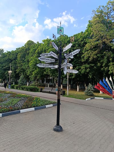 The image shows a signpost with multiple directional arrows in Gubkin, Russia. The signpost is black with white arrows pointing in various directions, each indicating a city and its distance. Some of the legible destinations include "Madrid" (336 km), "Keyptayn" (9656 km), "Rio-de-Janeiro" (11362 km), "Pekin" (5961 km), "Belgorod" (101 km), "Antalya" (6400 km), "Center" (912 km), "Rodinskoe Pole" (599 km), and "Solntse" (150 million km). The signpost is located in a park with trees, benches, a flower bed, and a paved walkway. Two people are walking in the background, and some colorful flags are visible on the right side of the image. The sky is blue with white clouds. The overall scene depicts a public park in Gubkin, Russia, featuring a notable landmark signpost.