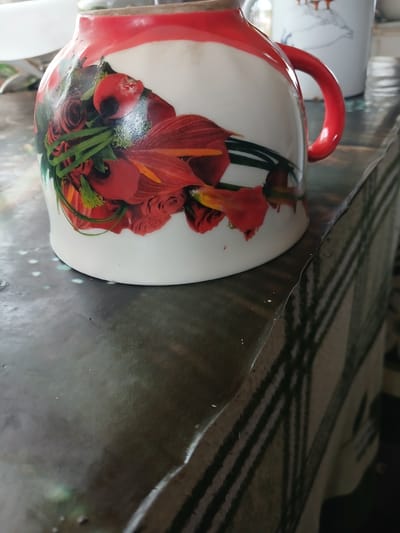 A close-up, low-angle shot shows a white ceramic pitcher with a red rim and a floral design of red flowers and green leaves. The pitcher has a red handle. It rests on a dark, reflective surface that appears to be a table covered with a cloth that has a green and white striped pattern. The background is blurred, showing another white mug with a duck illustration and a hint of a silver lid or container. The scene is indoors, possibly in a home environment in Hlushky, Ukraine, based on the location tag. There are no people visible in the image.